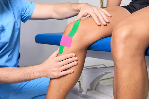 Therapist applies green and pink kinesiology tape to a patient's knee.
